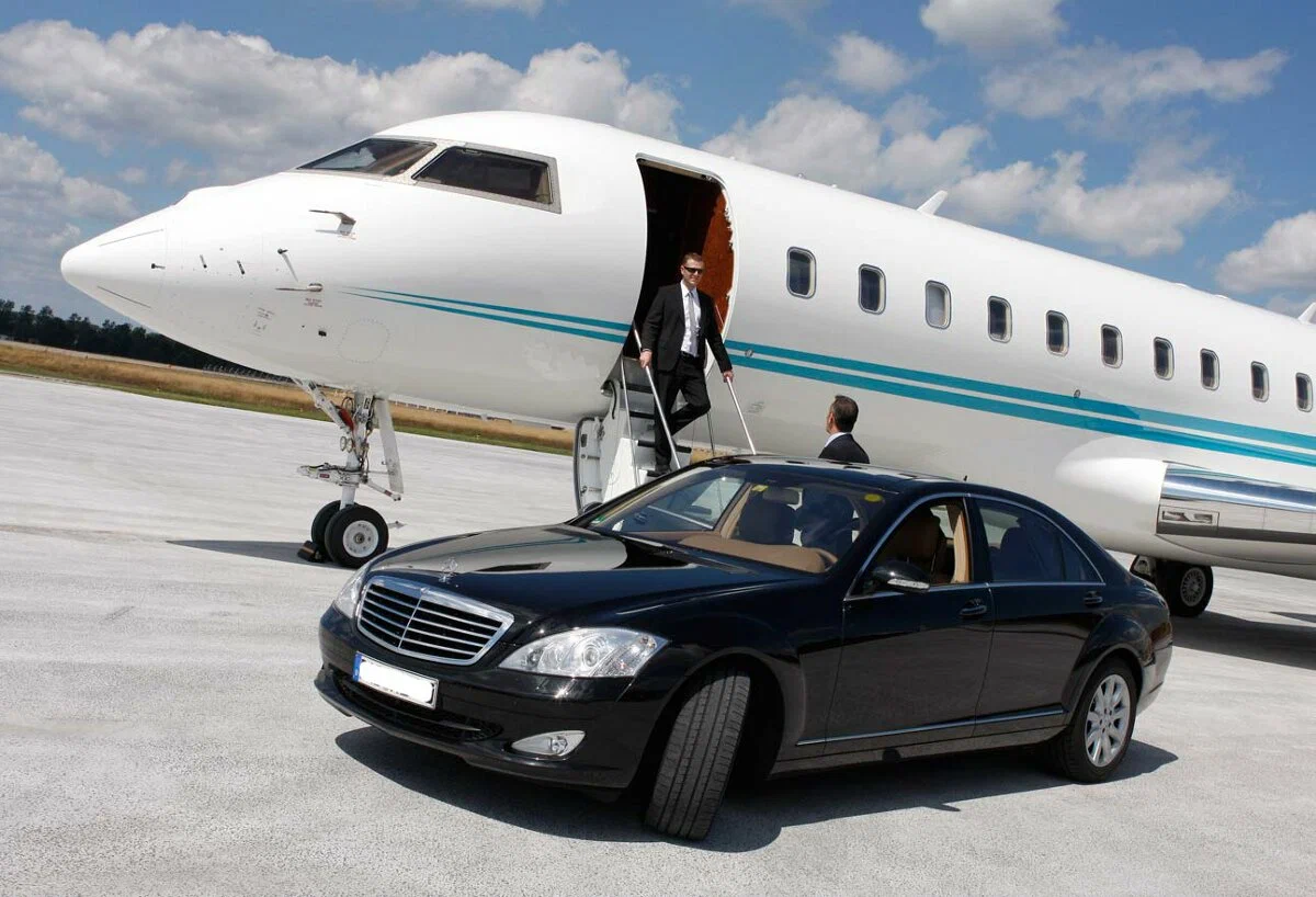 A man in a suit descends steps from a private jet toward a black luxury sedan parked on an airport tarmac under a partly cloudy sky for a client who uses the burlington limo service.