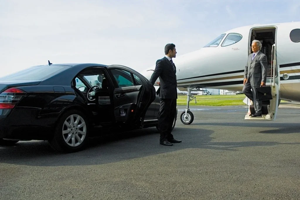 A businessman in a suit exits a private jet holding a briefcase, greeted by a chauffeur of burlington limo service beside a black luxury car with an open door on a sunny day.