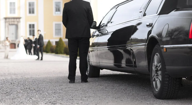 waterloo airport limo services-A suited man stands beside a black limousine, parked on gravel. In the background, a bride and groom pose with others near a yellow building, conveying a formal, celebratory mood.