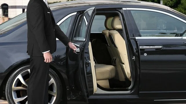 cambridge airport limo service-A well-dressed chauffeur opens the door of a luxury black car, revealing beige leather seats inside.