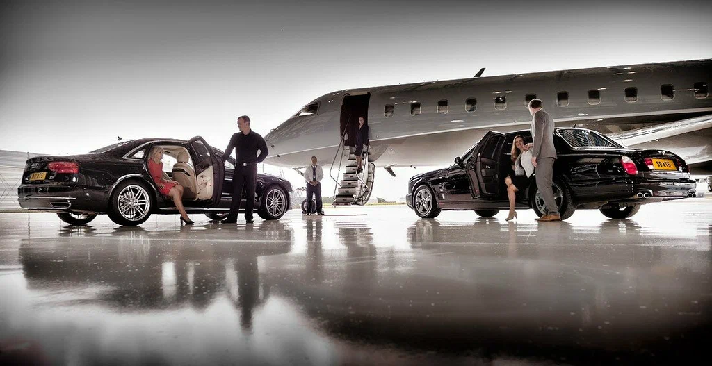 book a limo service-A luxurious scene at an airport hangar with two sleek cars, passengers in elegant attire, and a private jet in the background.
