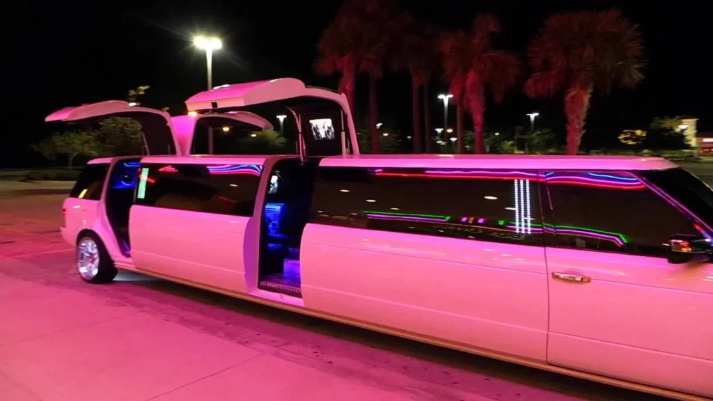 Brampton to pearson airport limo-A white stretch limousine with upward-opening doors is illuminated by pink and blue lights at night, parked near palm trees under streetlights.