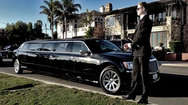 Alliston airport limo services-A sleek black limousine parked on a suburban street, with a chauffeur in a suit standing beside it. Palm trees and elegant houses are visible in the background.