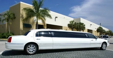 Aurora airport limo service-A long white limousine is parked on a cobblestone driveway in front of a modern building with palm trees, conveying a luxurious and sunny setting.