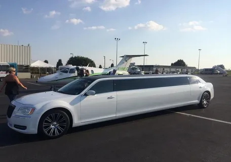 kingston limo service-A long white limousine parked at an airport with small planes in the background. A few people are nearby on the sunny tarmac, conveying a sense of luxury.