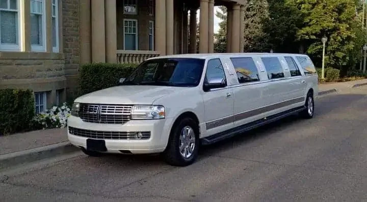limo service orangeville-A long white limousine is parked on a quiet street beside a stately building with large columns. The surroundings are lush and green, conveying luxury and elegance.
