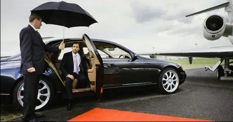 markham limo service-A man in a suit exits a black luxury car onto a red carpet as another man holds an umbrella. A private jet is parked nearby under a cloudy sky.