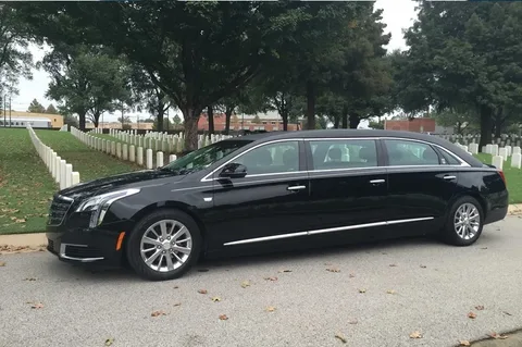 A sleek black limousine is parked on a road beside a cemetery with white headstones. The scene is calm and somber, under a cloudy sky.