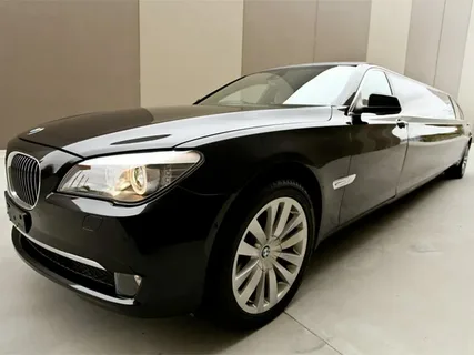 A sleek black limousine is parked indoors, angled to show its elongated body. The luxury vehicle has shiny alloy wheels and striking headlights.