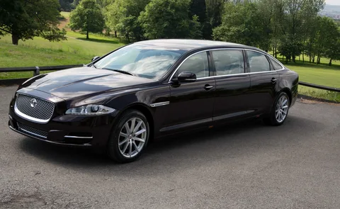 A sleek black luxury sedan is parked on a paved road, surrounded by lush green trees and grass under a clear sky, conveying elegance and tranquility.