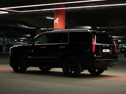A black SUV is parked in a dimly lit underground garage. The vehicle's sleek design stands out against the red pillars and concrete surroundings, creating a mysterious, urban atmosphere.