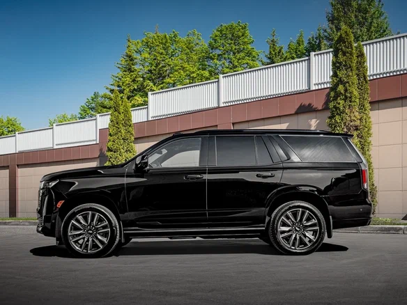 A sleek black SUV is parked on a sunny day, showcasing a glossy finish. In the background, there are trees and a modern fence, creating a polished and stylish scene.