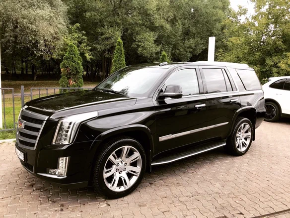 A sleek black SUV parked on a paved area, framed by lush green trees. The vehicle is polished, reflecting light, and exudes a sense of luxury and power.