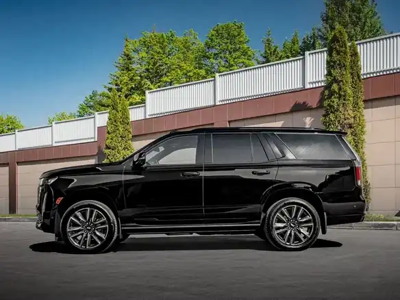 A sleek black SUV is parked on a sunny day, showcasing a glossy finish. In the background, there are trees and a modern fence, creating a polished and stylish scene.