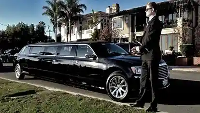 A sleek black limousine parked on a suburban street, with a chauffeur in a suit standing beside it. Palm trees and elegant houses are visible in the background.