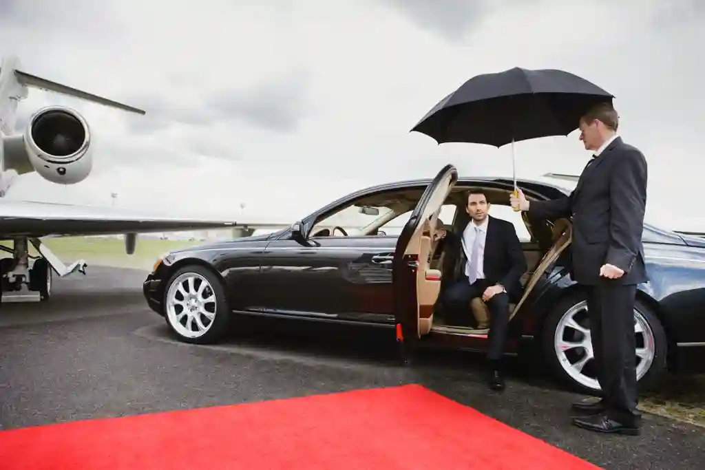 A luxury black car parked on a red carpet near a private jet, with a man holding an umbrella for another person seated inside.